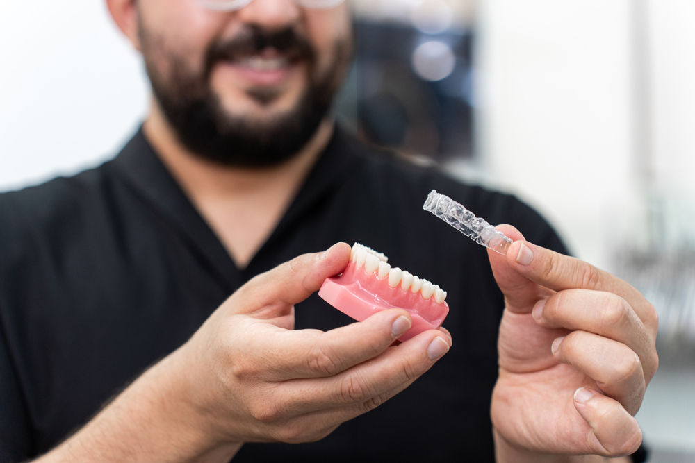 Smiling doctor installing clear aligners on plastic human jaw model presenting teeth correction
